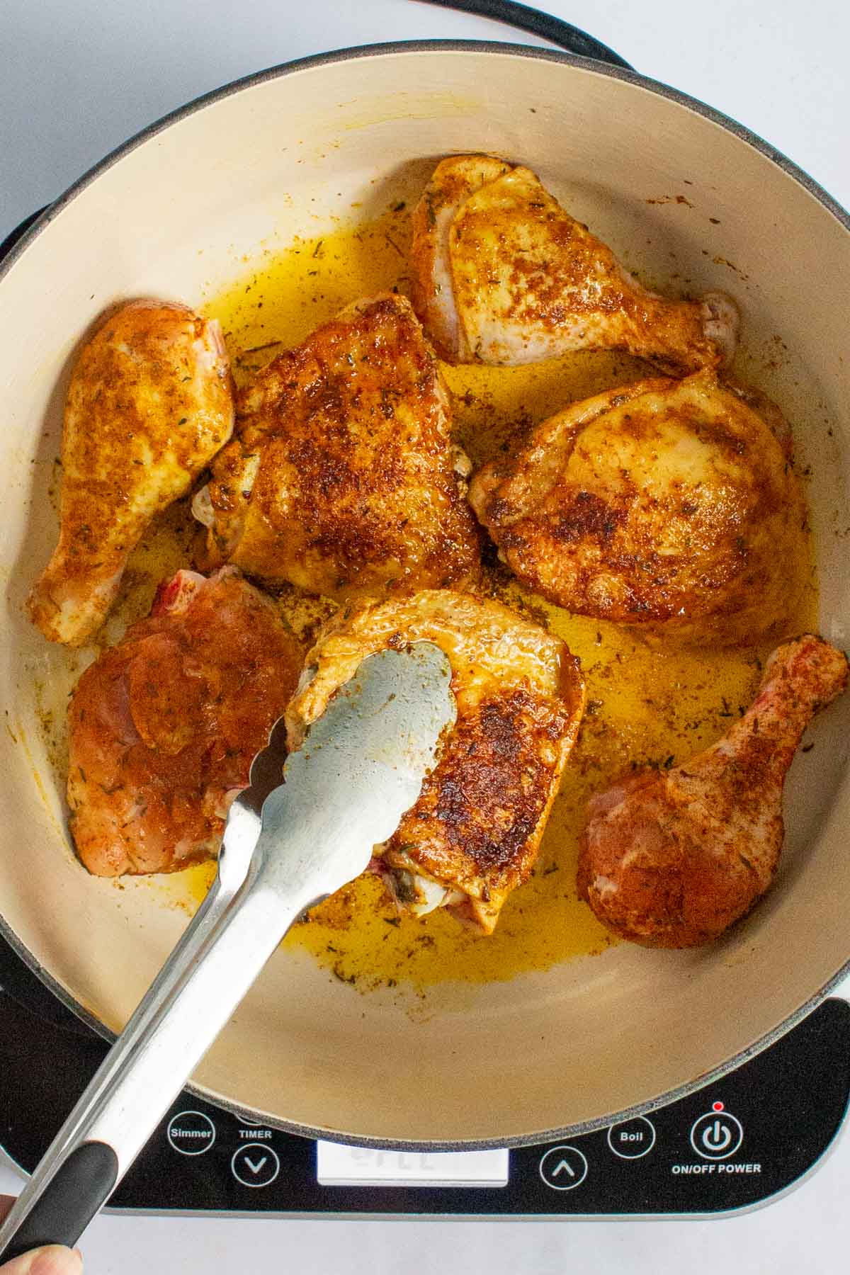 Tongs holding up a piece of browned chicken in Dutch oven for Nigerian Red Stew.