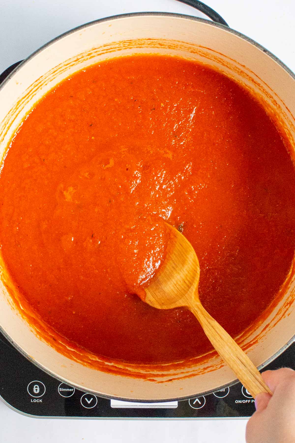 A hand using a wooden spoon to stir sauce in a Dutch oven for Nigerian Red Stew.