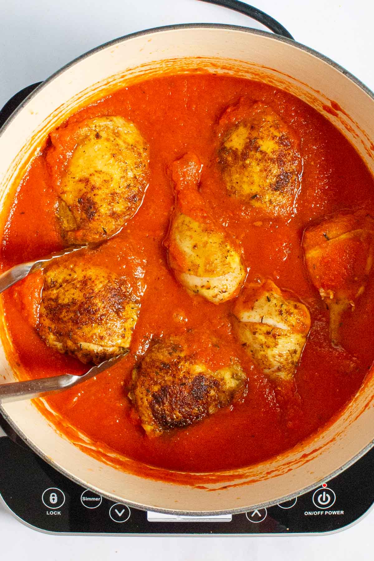 Tongs nestling browned chicken pieces into sauce for Nigerian Red Stew.