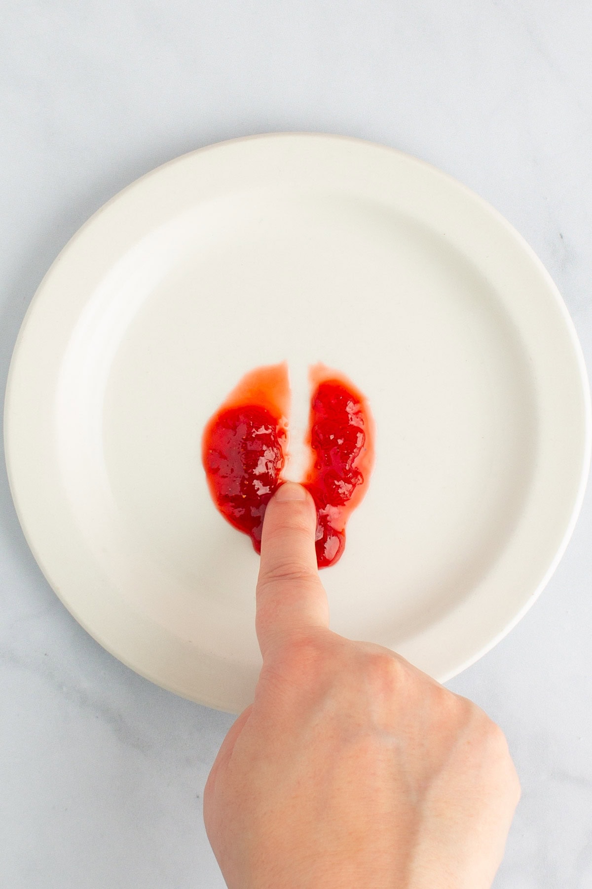 A finger drawing a line through a dollop of strawberry rhubarb jam on a plate to test the set.