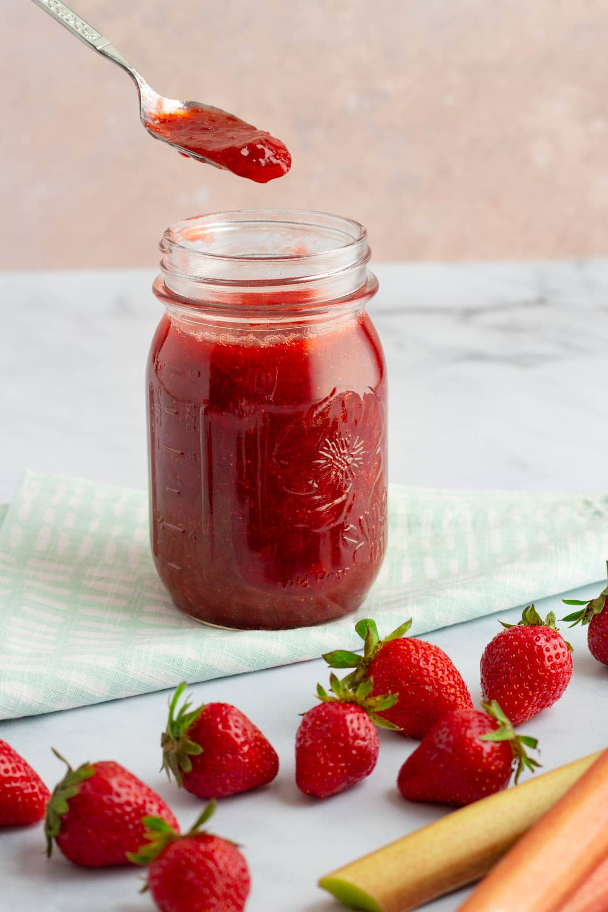 A small spoon over a jar filled with simple small-batch strawberry rhubarb jam, with strawberries and rhubarb on the counter.