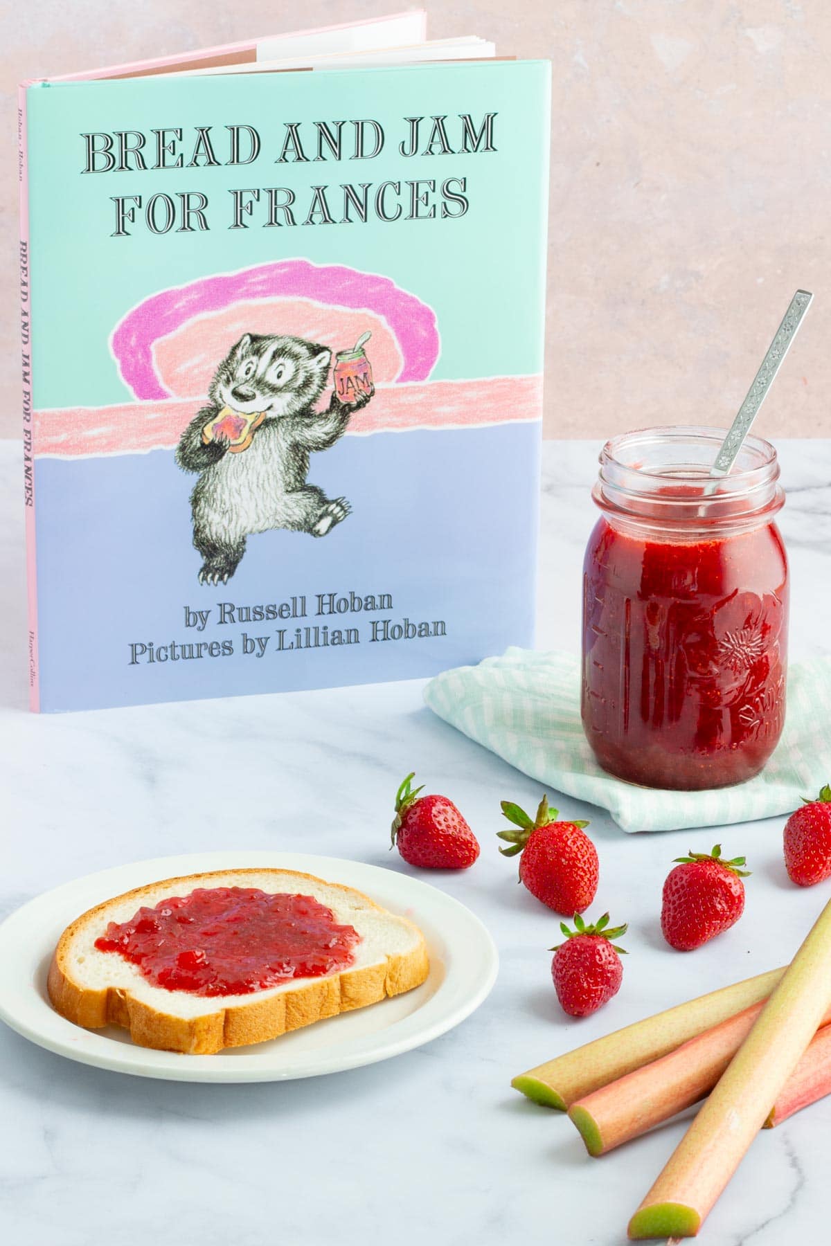 The book "Bread and Jam for Frances" next to a jar of simple small-batch strawberry rhubarb jam, a piece of bread spread with jam on a plate, and strawberries and rhubarb on the counter.