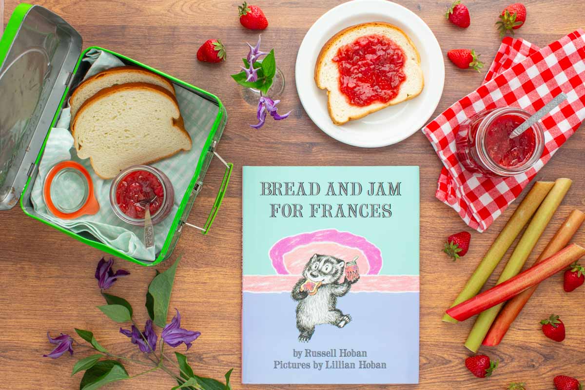 The book "Bread and Jam For Frances" next to a jar of strawberry rhubarb jam, a plate with a piece of bread with jam on it, an open lunchbox with more bread and jam in it, and strawberries and rhubarb scattered over the surface.