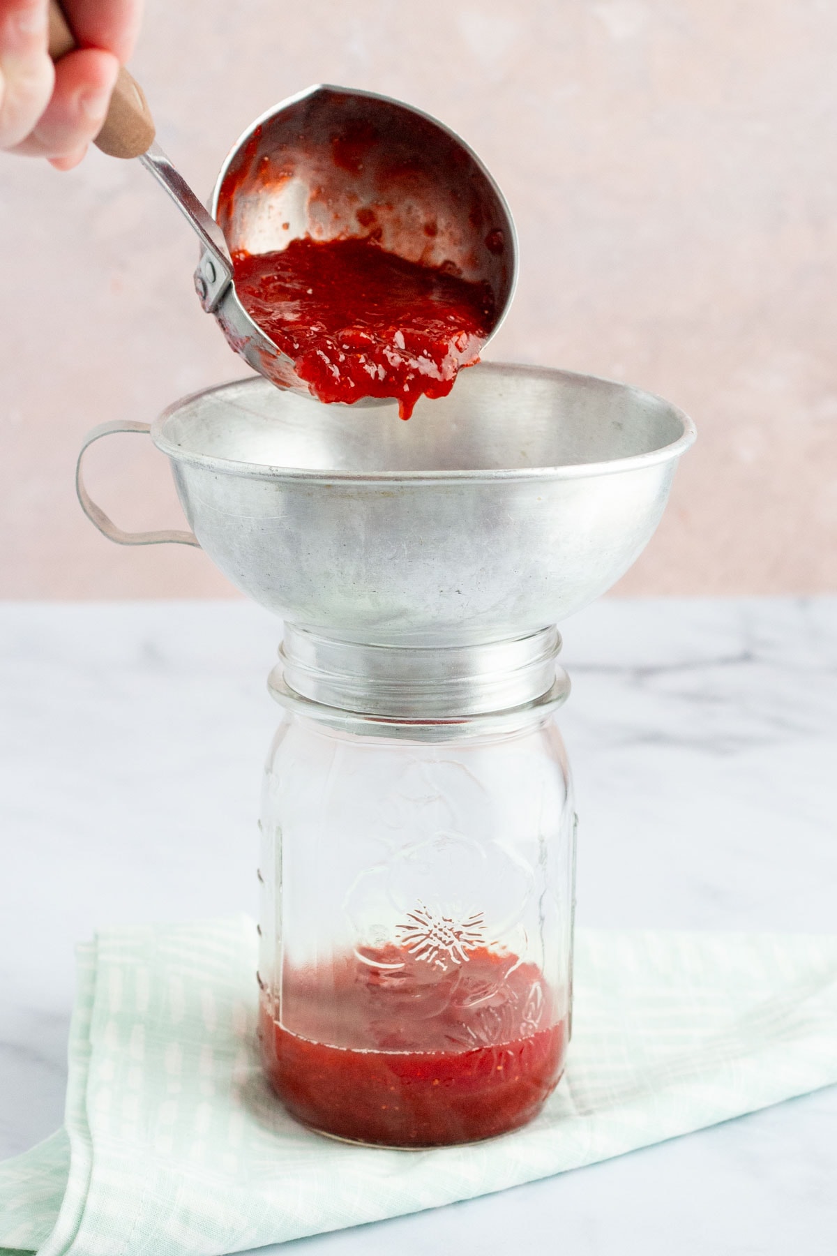 A hand using a ladle to transfer cooked strawberry rhubarb jam to a mason jar.