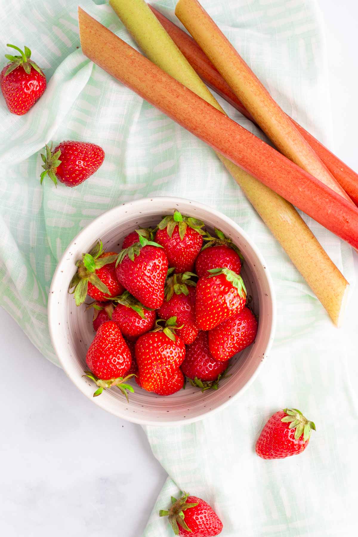 A bowl of fresh strawberries next to stalks of fresh rhubarb.