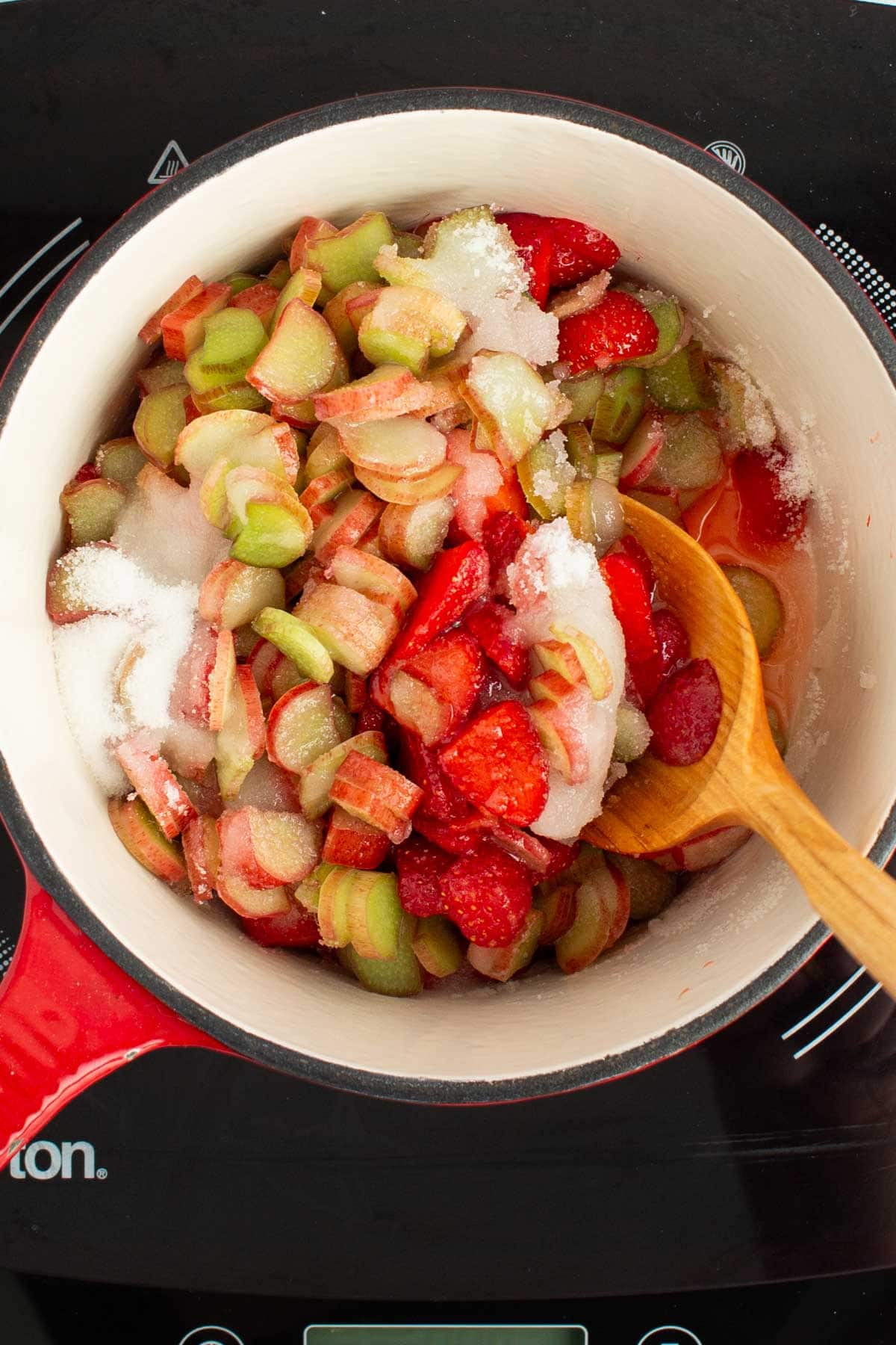 A spoon stirring together ingredients for strawberry rhubarb jam in a saucepan.