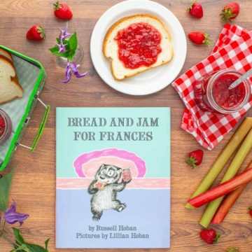 The book "Bread and Jam For Frances" next to a jar of strawberry rhubarb jam, a plate with a piece of bread with jam on it, an open lunchbox with more bread and jam in it, and strawberries and rhubarb scattered over the surface.