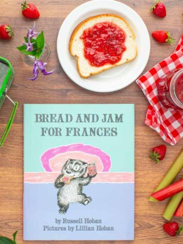 The book "Bread and Jam For Frances" next to a jar of strawberry rhubarb jam, a plate with a piece of bread with jam on it, an open lunchbox with more bread and jam in it, and strawberries and rhubarb scattered over the surface.