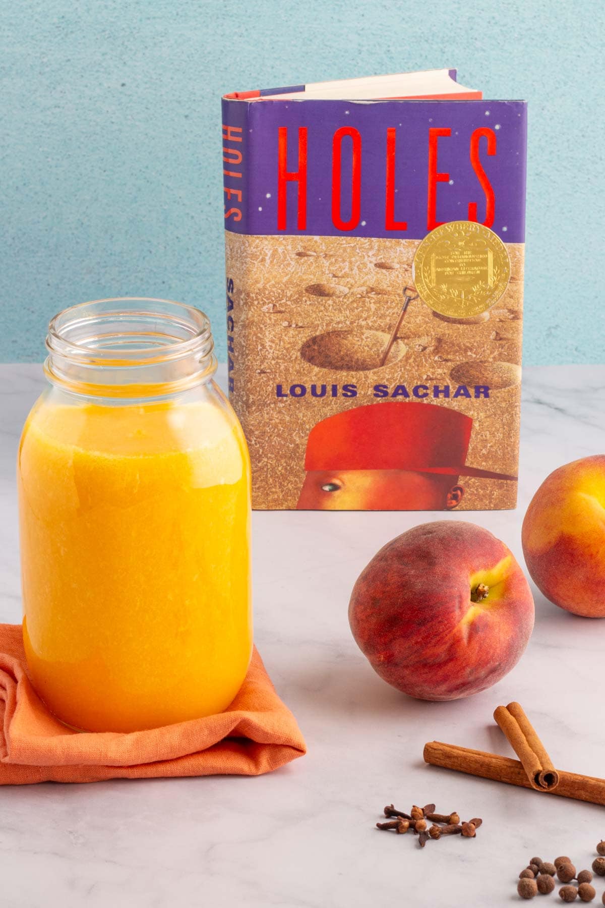 The cover of the book Holes with a jar of spiced peach nectar, some fresh peaches, and some whole spices next to it.