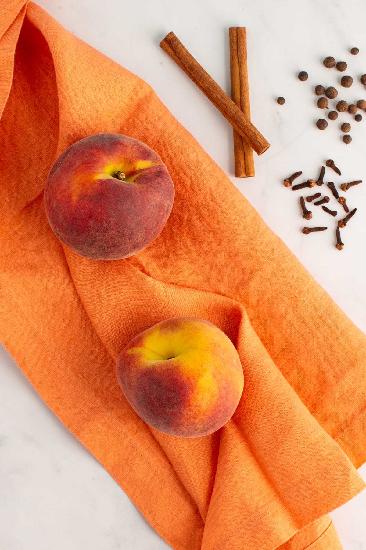 Fresh peaches on a towel, with cinnamon sticks, whole cloves, and allspice berries nearby.