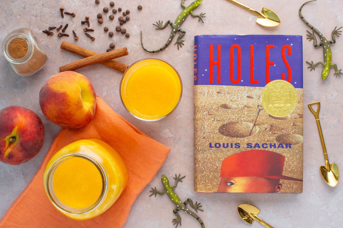 The cover of the book Holes along with a mason jar and drinking glass filled with spiced peach nectar, with fresh peaches, spices, toy lizards, and small shovels next to them on a surface.