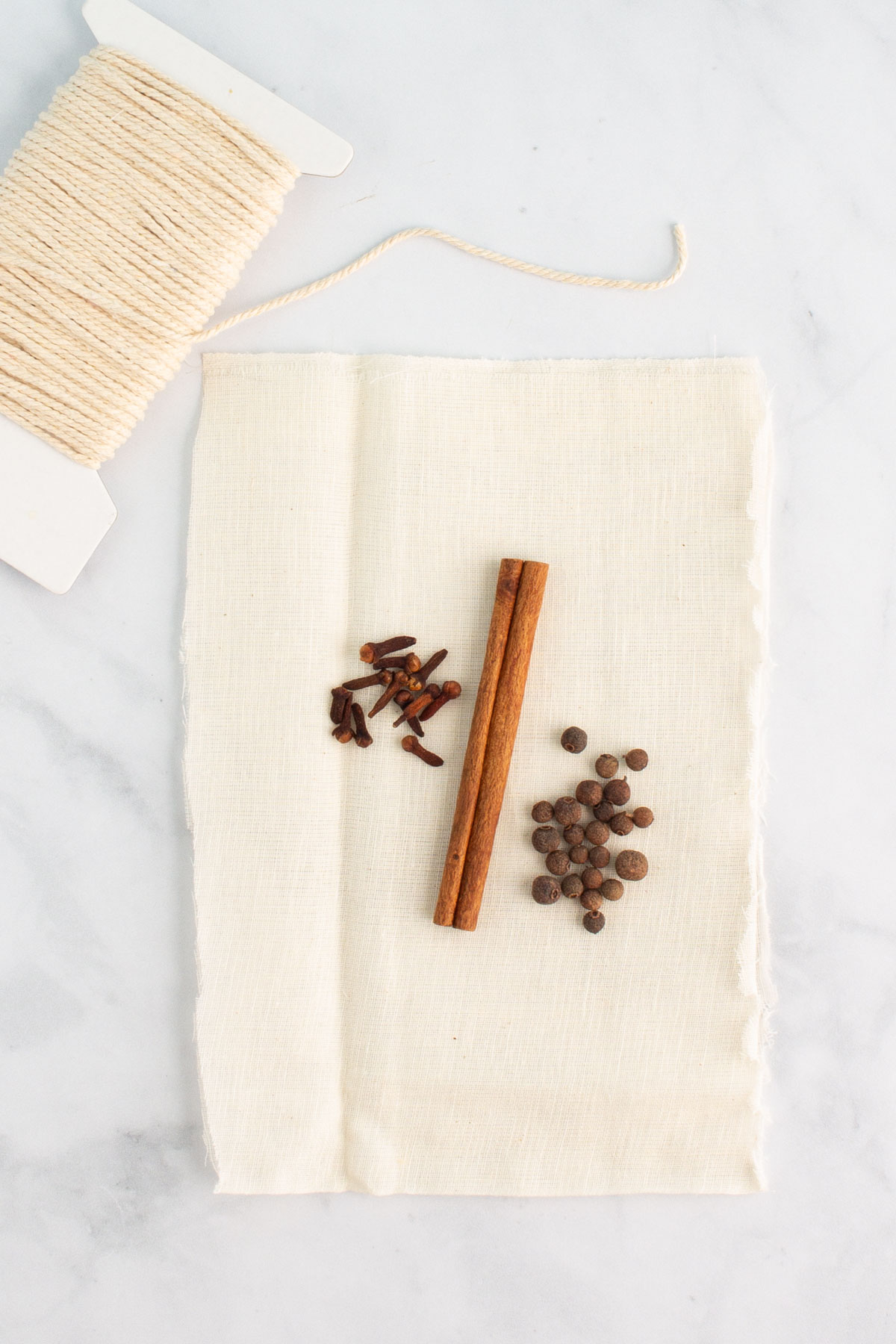 A cinnamon stick, whole cloves, and allspice berries on a piece of cheesecloth, with kitchen twine nearby.