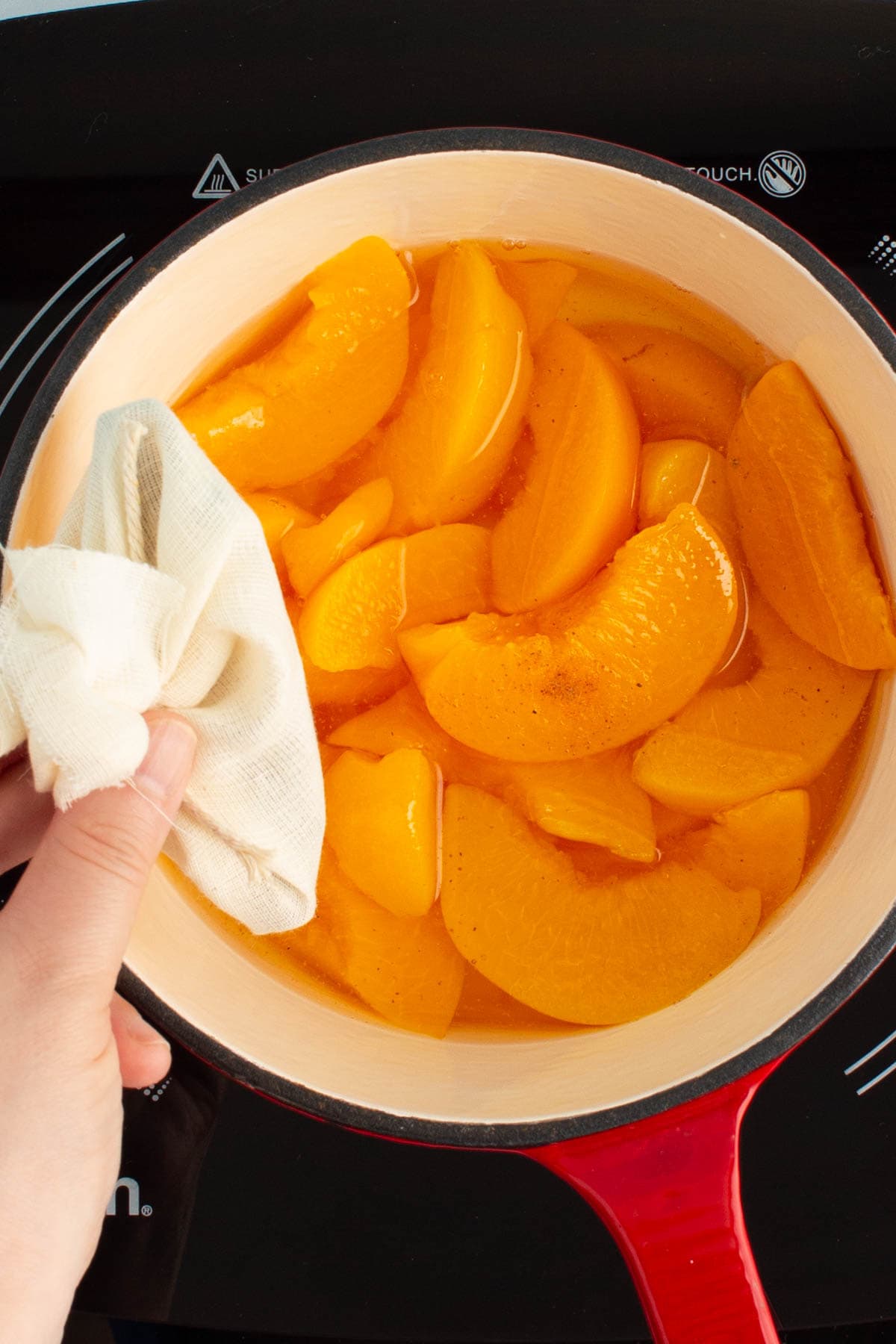 A hand adding a sachet of spices to a saucepan of sliced peaches for spiced peach nectar.