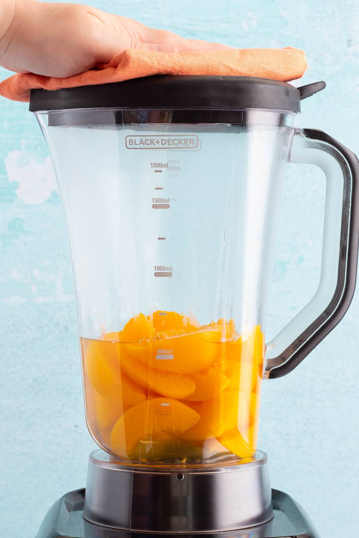 A hand holding the lid on a blender with a peach mixture inside.