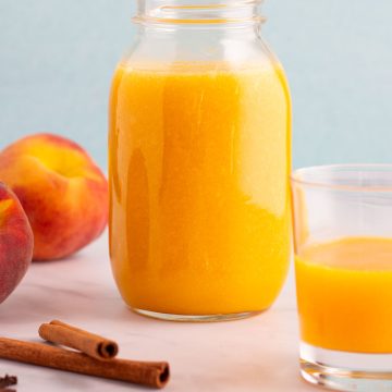 A mason jar and glass filled with spiced peach nectar, with spices and peaches on the counter nearby.