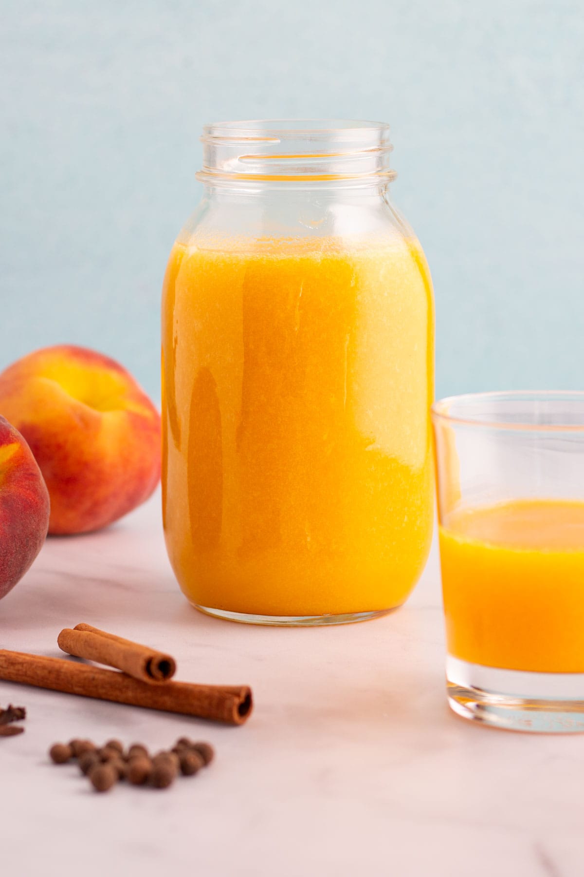 A mason jar and glass filled with spiced peach nectar, with spices and peaches on the counter nearby.