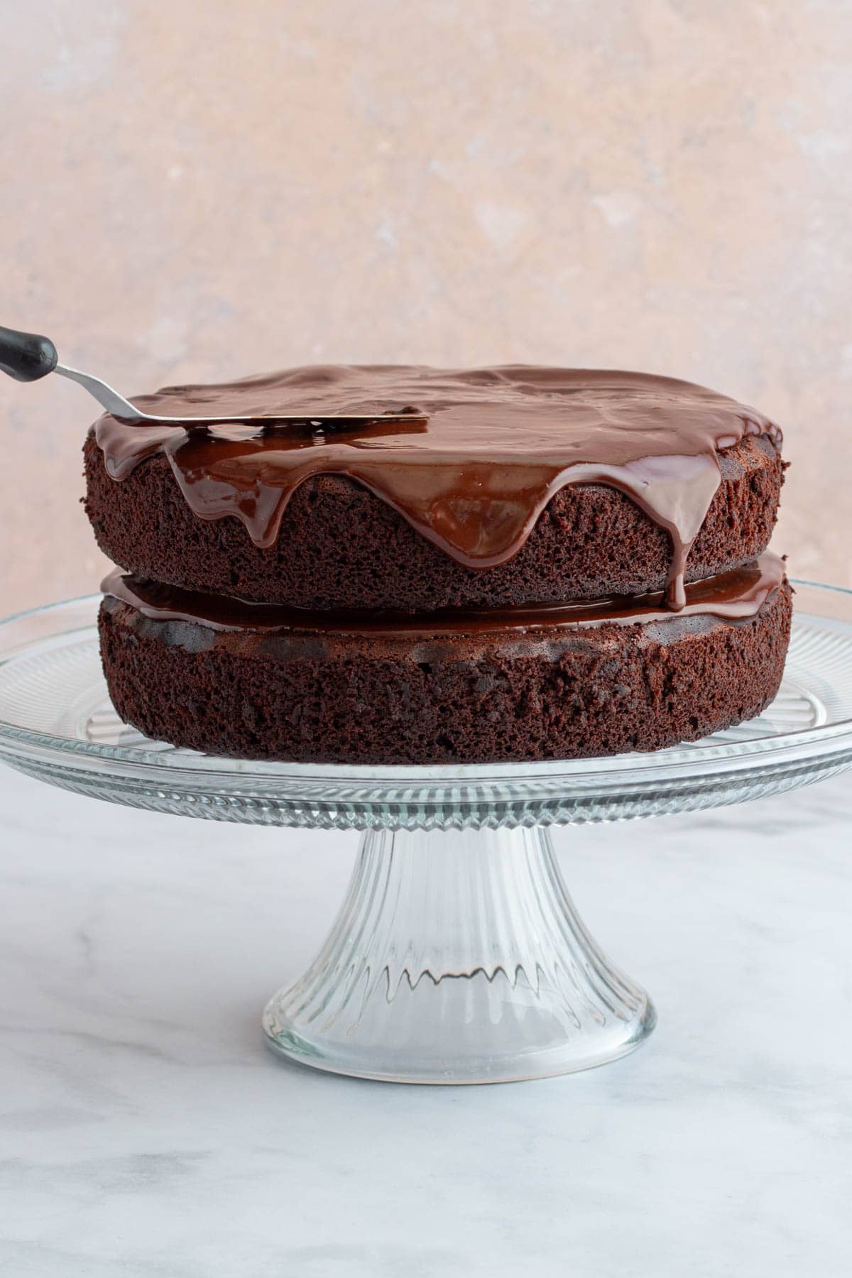 An offset spatula spreading melted chocolate ganache over the top of a chocolate ganache cake on a cake stand.