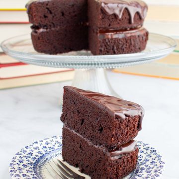 A slice of chocolate ganache cake on a plate with a fork, with the rest of the cake on a stand in the background.