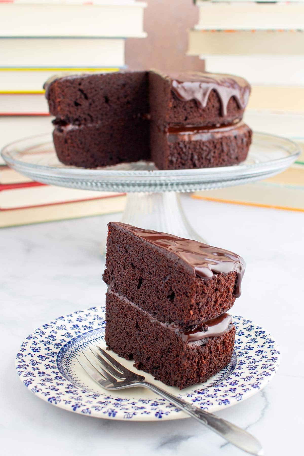 A slice of chocolate ganache cake on a plate with a fork, with the rest of the cake on a stand in the background.