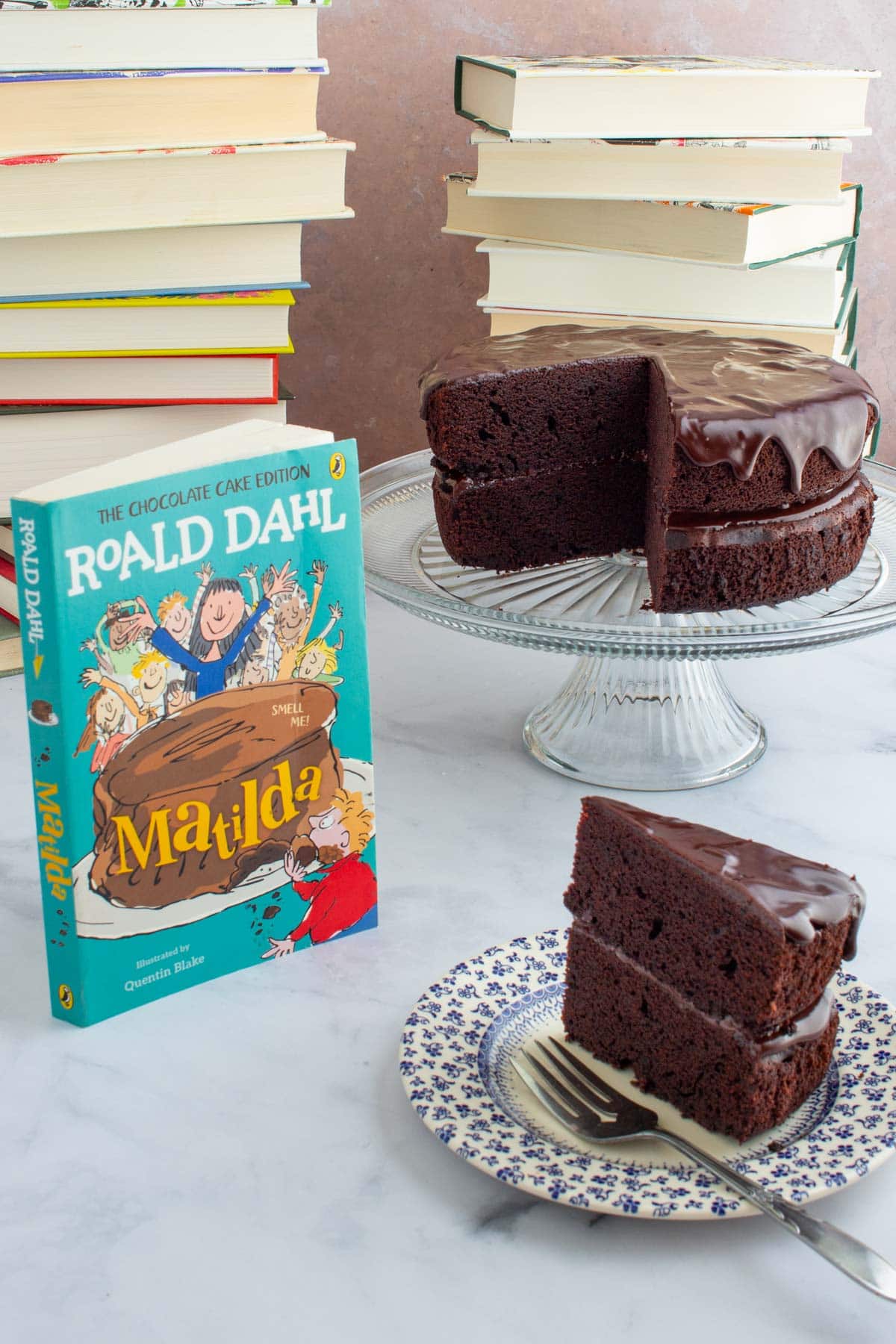 A slice of chocolate ganache cake on a plate with a fork next to the cover of the book Matilda, with the rest of the cake on a cake stand in the background.