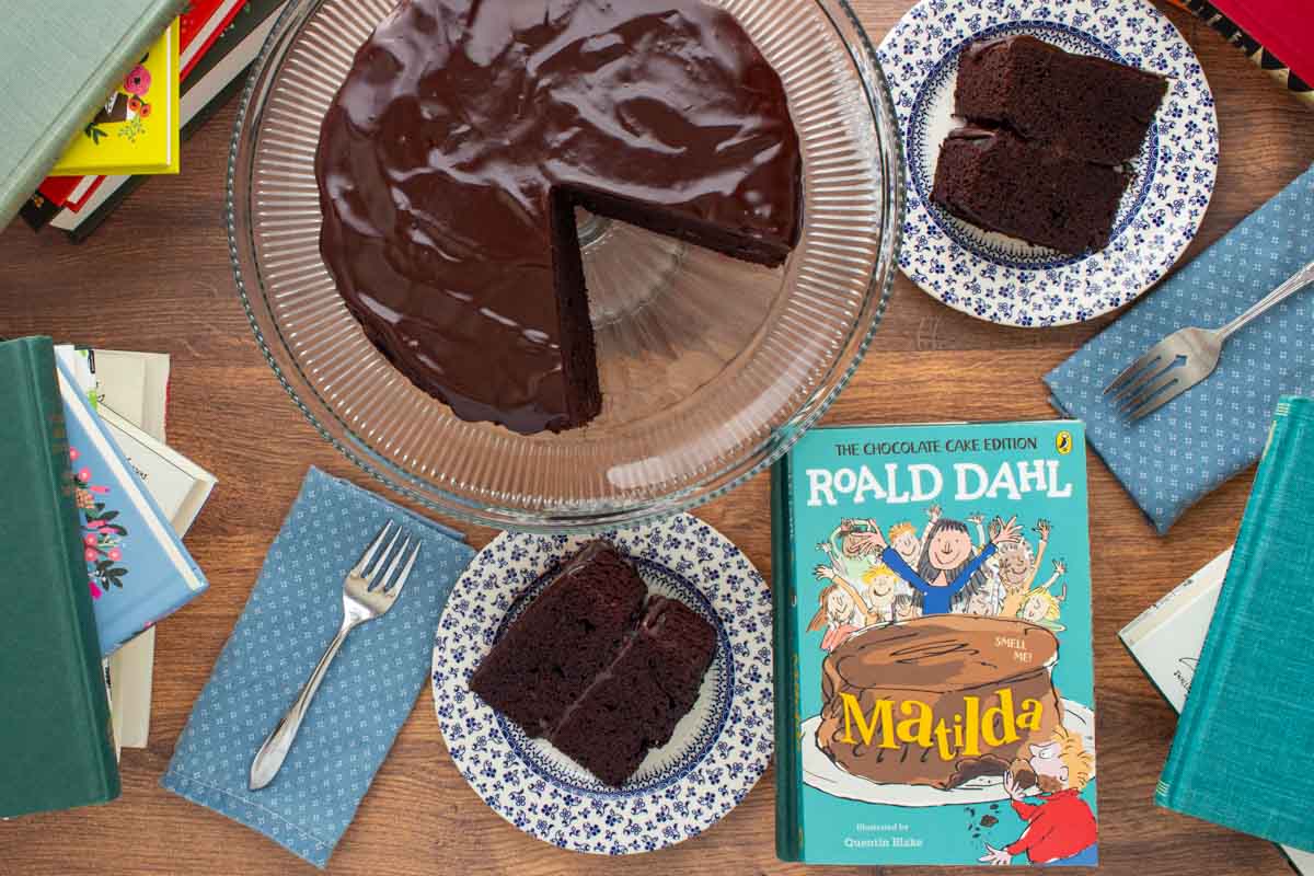 The cover of the book Matilda next to a chocolate ganache cake with a two slices cut out of it, with cake slices on small plates with forks, and stacks of books at the edges.