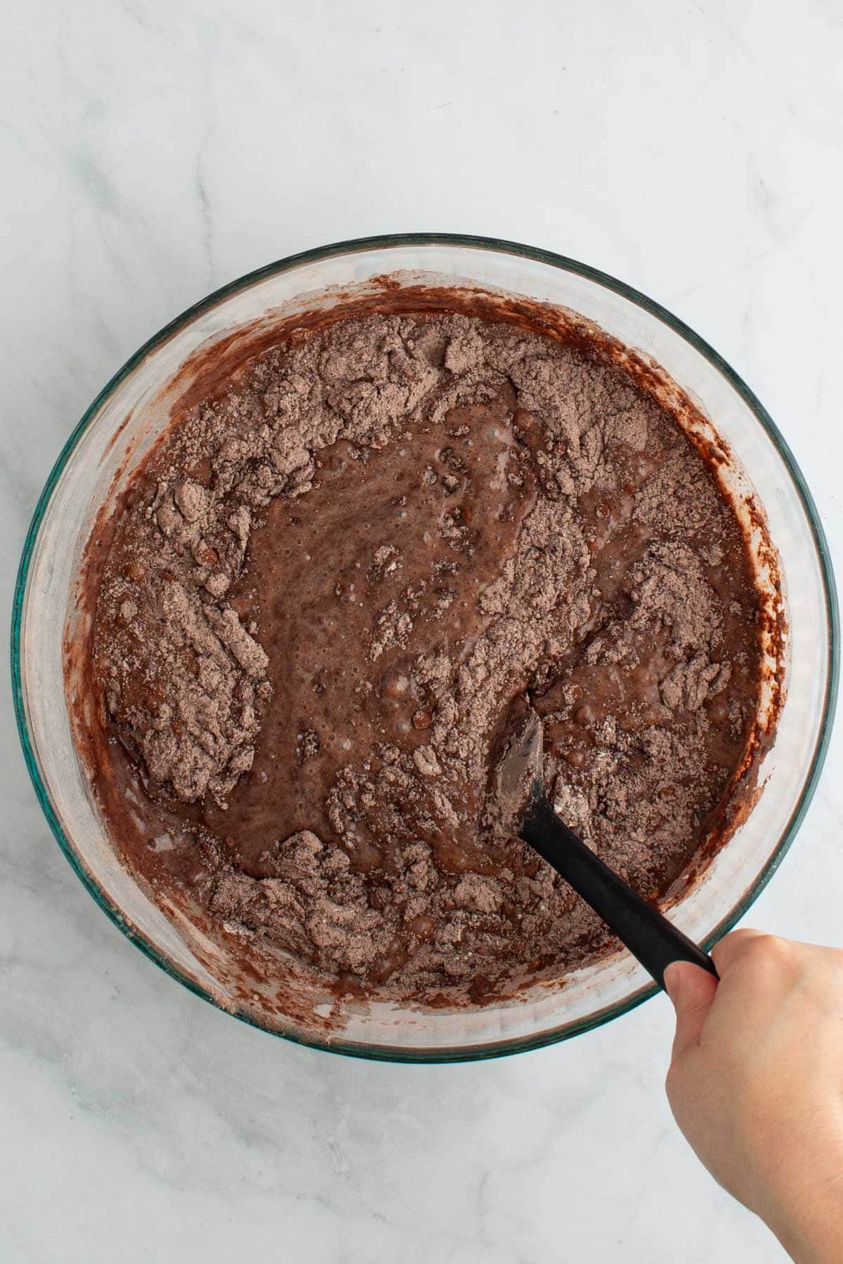 A hand using a rubber spatula to stir together batter for chocolate ganache cake in a bowl.