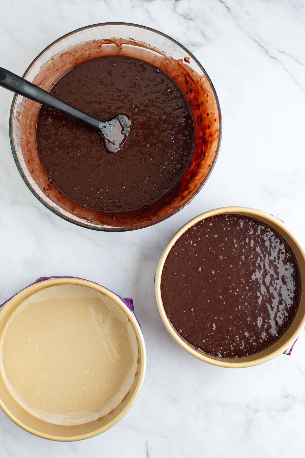 A bowl with chocolate ganache cake batter next to one filled cake pan, with an empty cake pan next to it ready to fill.