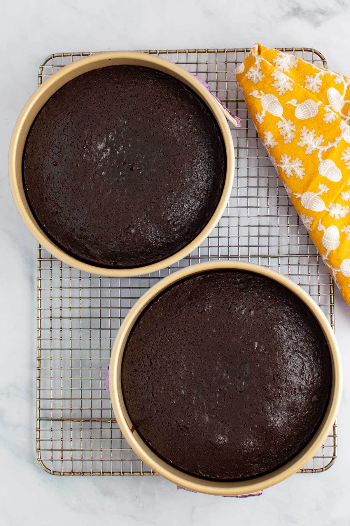 Two baked layers of chocolate ganache cake in pans on a cooling rack.