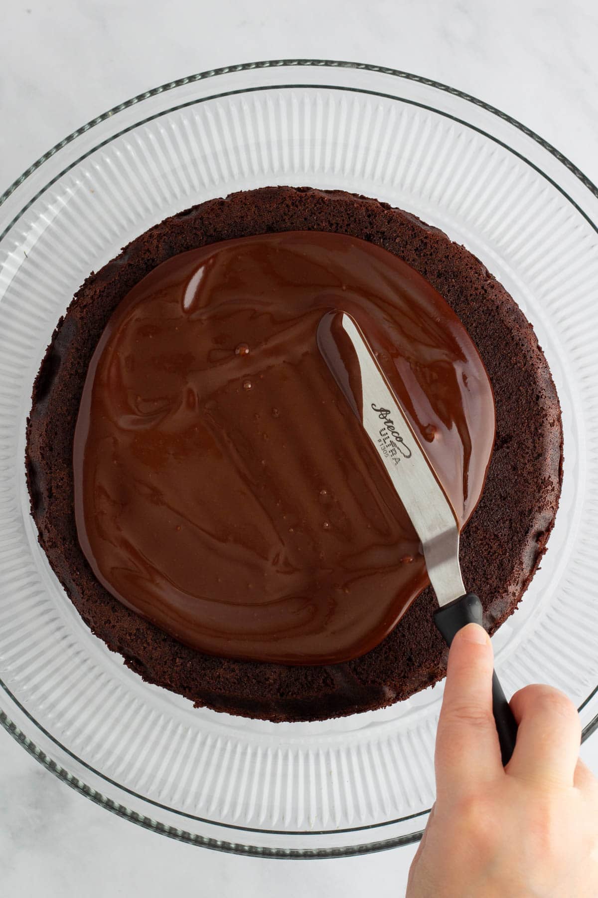 A hand using a small offset spatula to spread chocolate ganache on a cake layer.