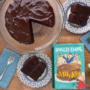 The cover of the book Matilda next to a chocolate ganache cake with a two slices cut out of it, with cake slices on small plates with forks.