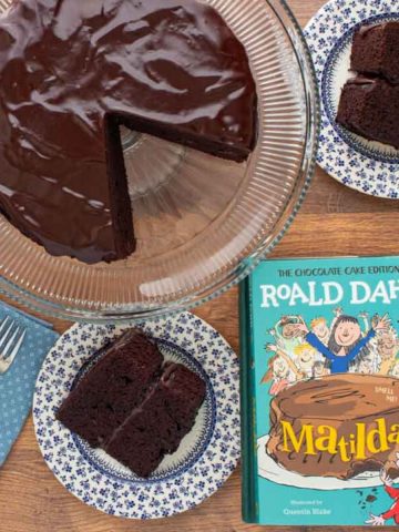 The cover of the book Matilda next to a chocolate ganache cake with a two slices cut out of it, with cake slices on small plates with forks.