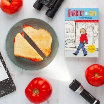 The cover of the book Harriet the Spy next to a tomato sandwich on a plate, surrounded by three tomatoes, a flashlight, binoculars, and a notebook.