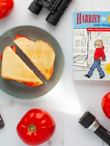 The cover of the book Harriet the Spy next to a tomato sandwich on a plate, surrounded by three tomatoes, a flashlight, binoculars, and a notebook.