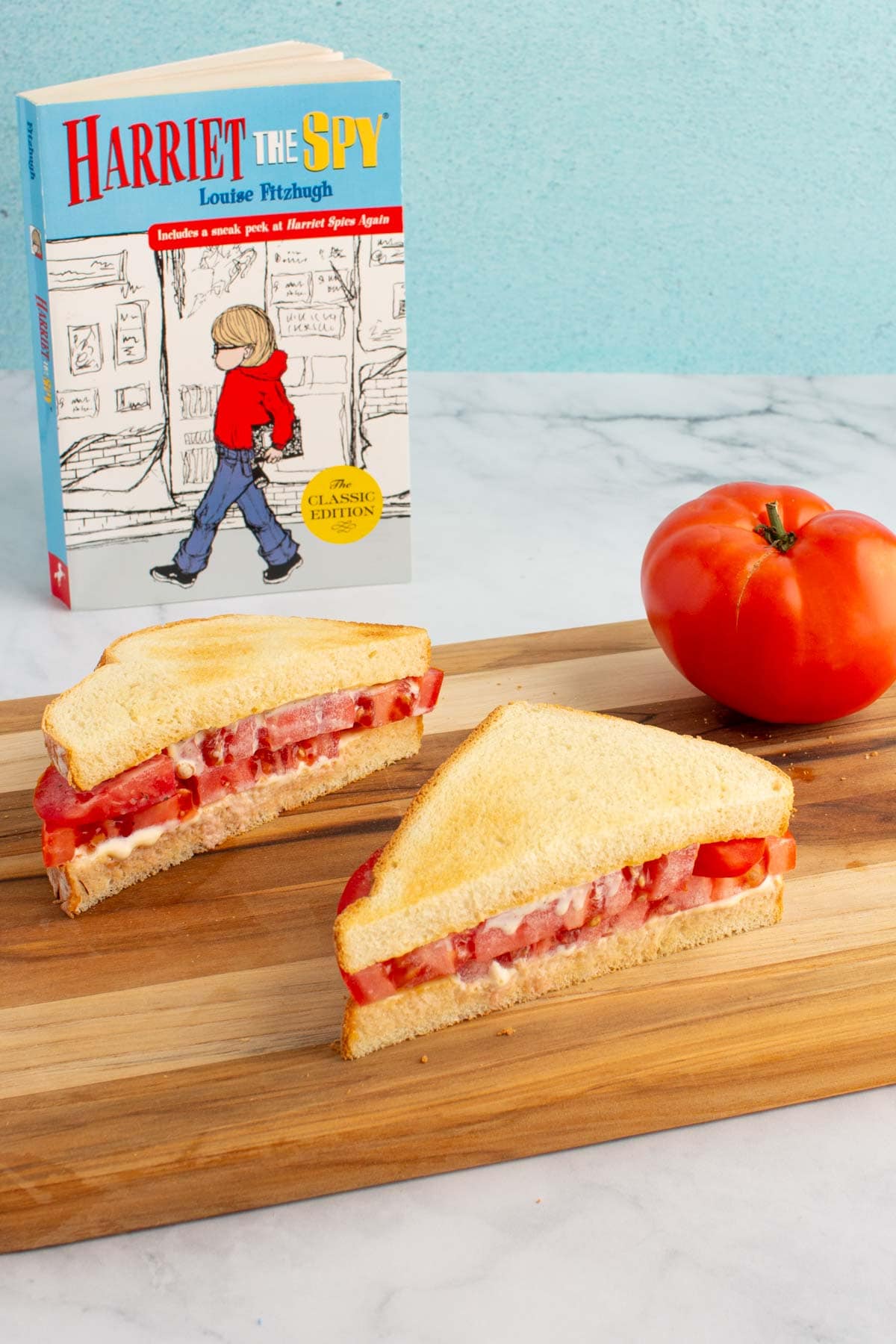 An ultimate umami tomato sandwich cut in half on a cutting board, with a tomato and the cover of the book Harriet the Spy nearby.