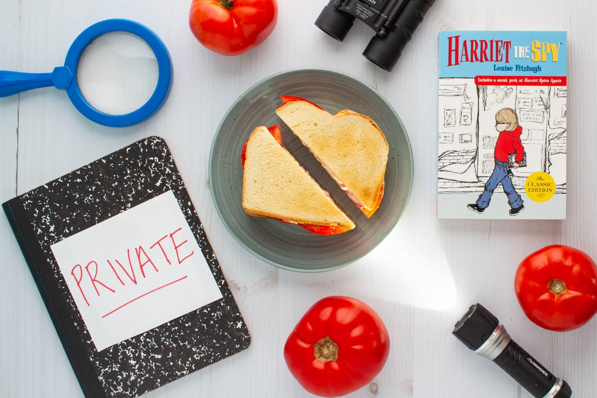 The cover of the book Harriet the Spy next to a tomato sandwich on a plate, surrounded by three tomatoes, a flashlight, binoculars, a magnifying glass, and a notebook.