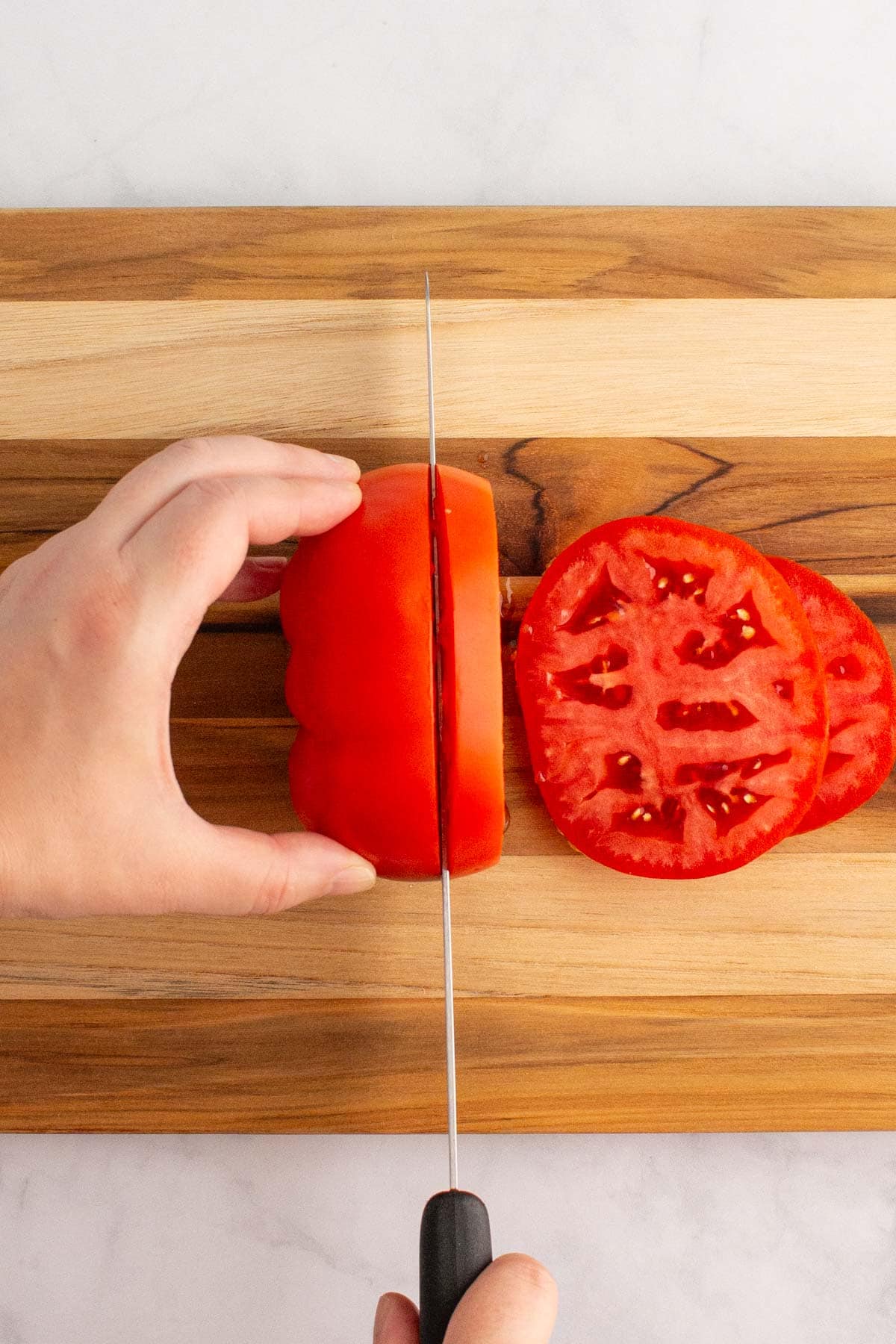 A hand cutting a tomato into slices on a cutting board.