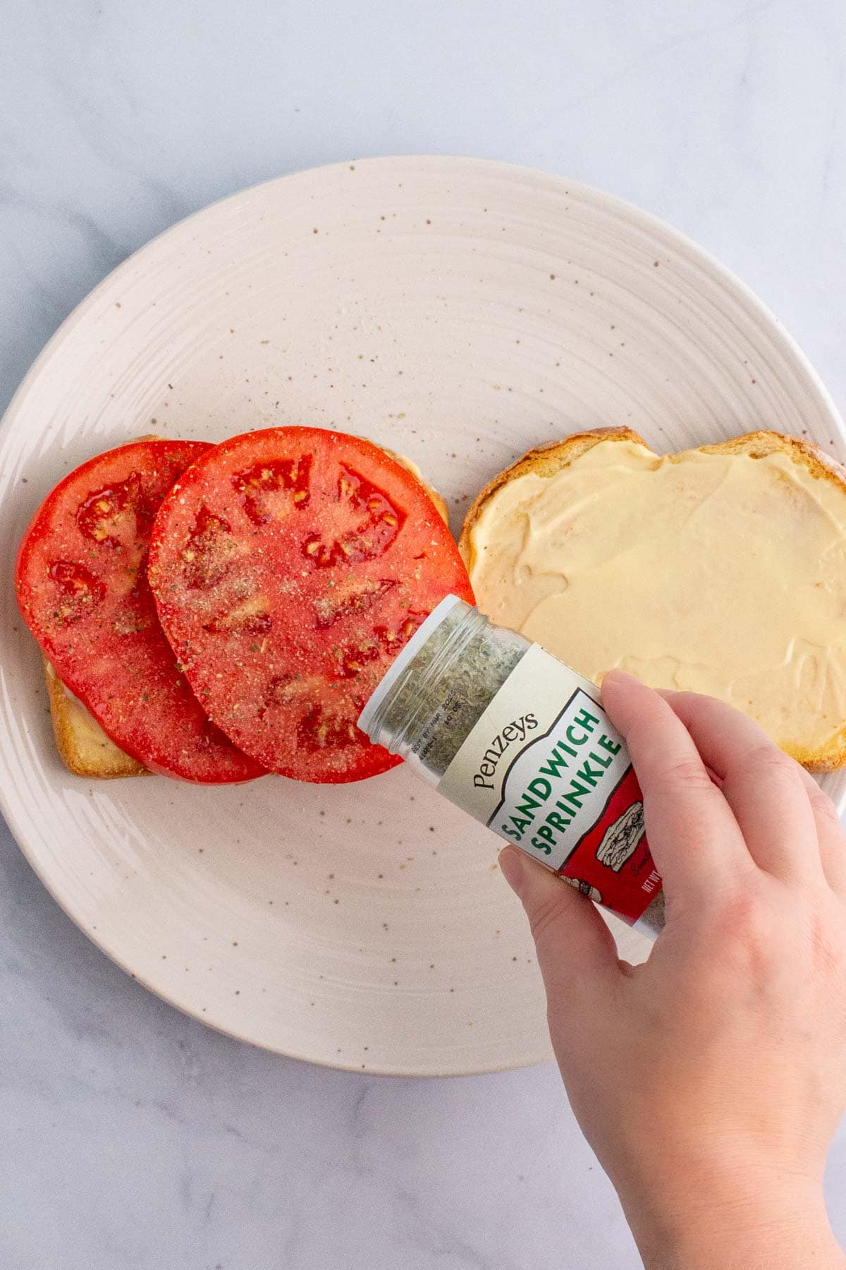 A hand shaking sandwich sprinkle seasoning onto tomatoes for an ultimate umami tomato sandwich.