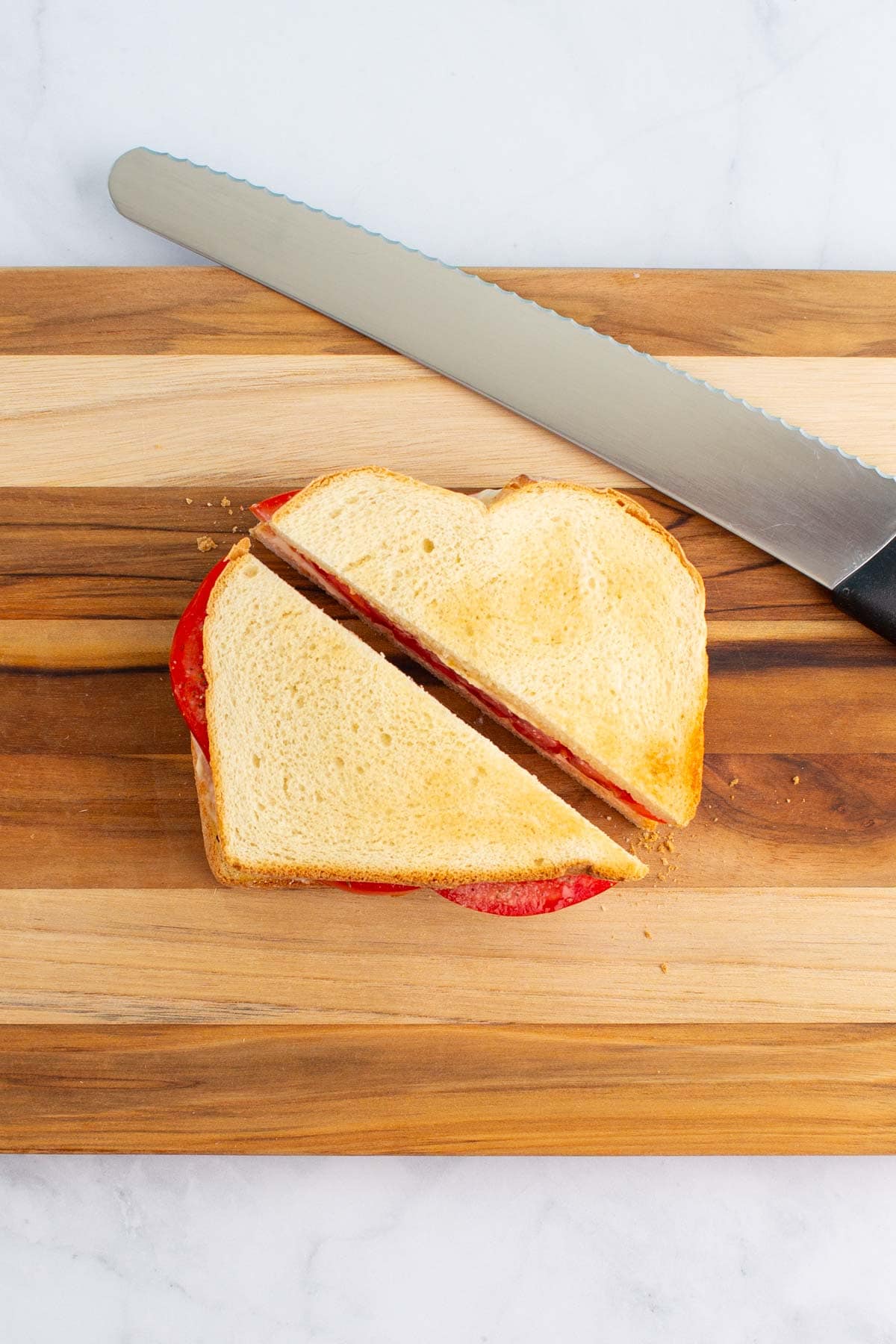 An ultimate umami tomato sandwich cut in half on a cutting board with a bread knife nearby.