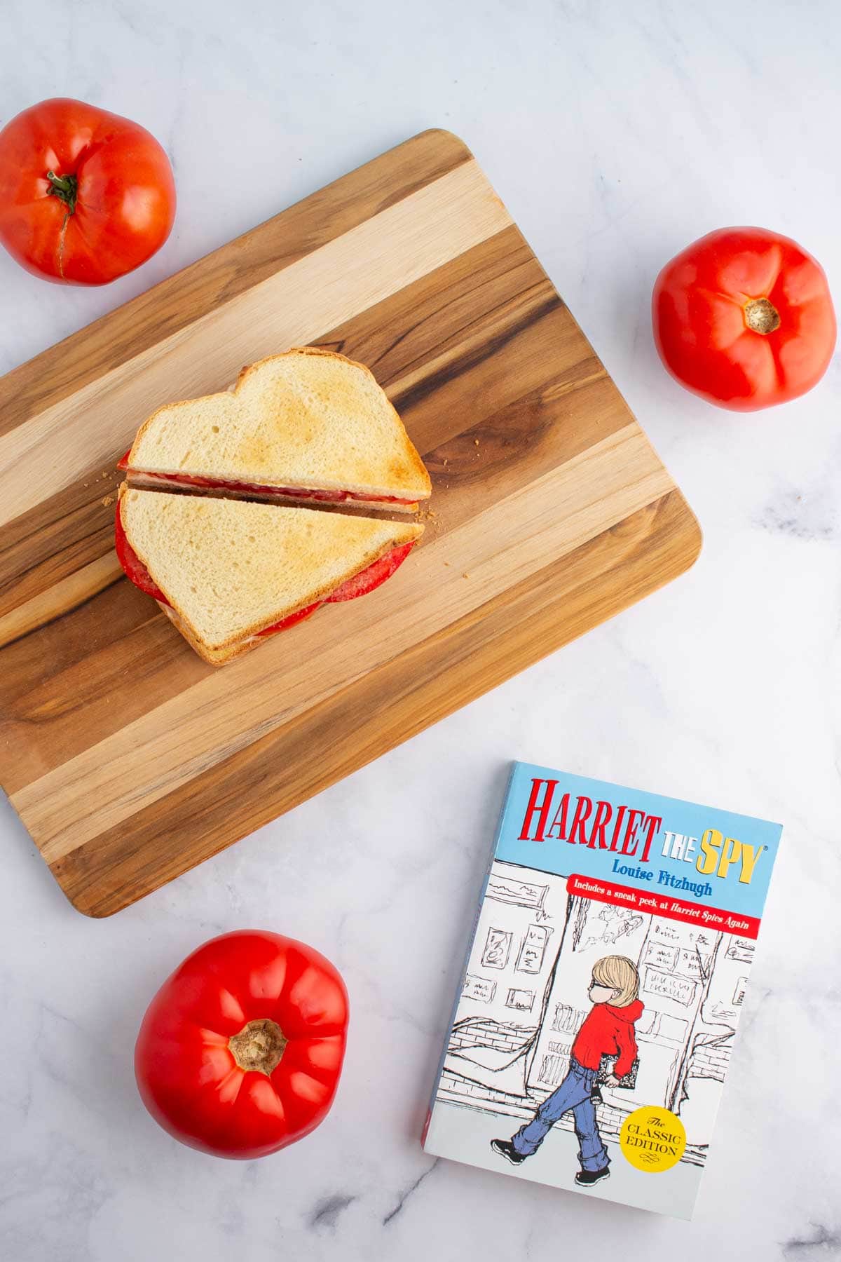 An ultimate umami tomato sandwich cut in half on a cutting board, with three tomatoes and the cover of the book Harriet the Spy nearby.