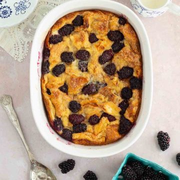 A baking dish of blackberry bread pudding with with fresh blackberries and a spoon next to it.