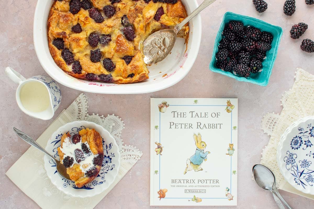 A baking dish of Blackberry Bread Pudding with the book The Tale of Peter Rabbit next to it, along with fresh blackberries, a spoon, and a pitcher of cream.