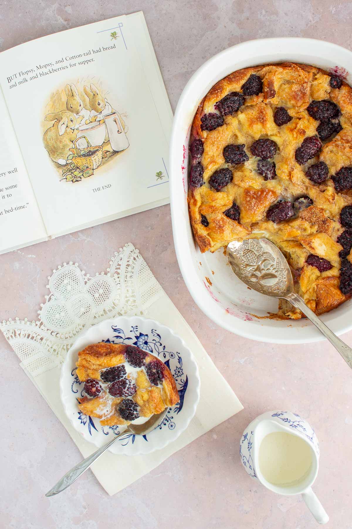 A baking dish of Blackberry Bread Pudding with the book The Tale of Peter Rabbit next to it open to a page, along with a serving of bread pudding in a dish and a pitcher of cream.