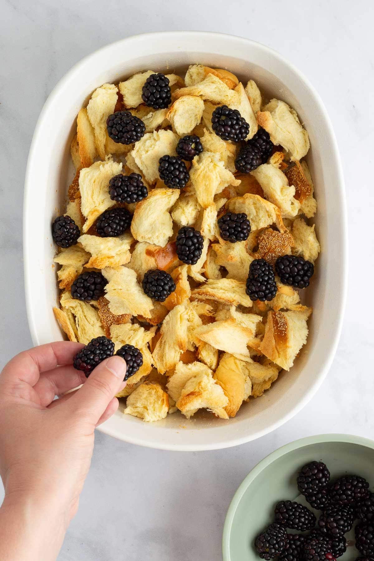 Toasted challah in a baking dish, with a hand adding fresh blackberries on top.