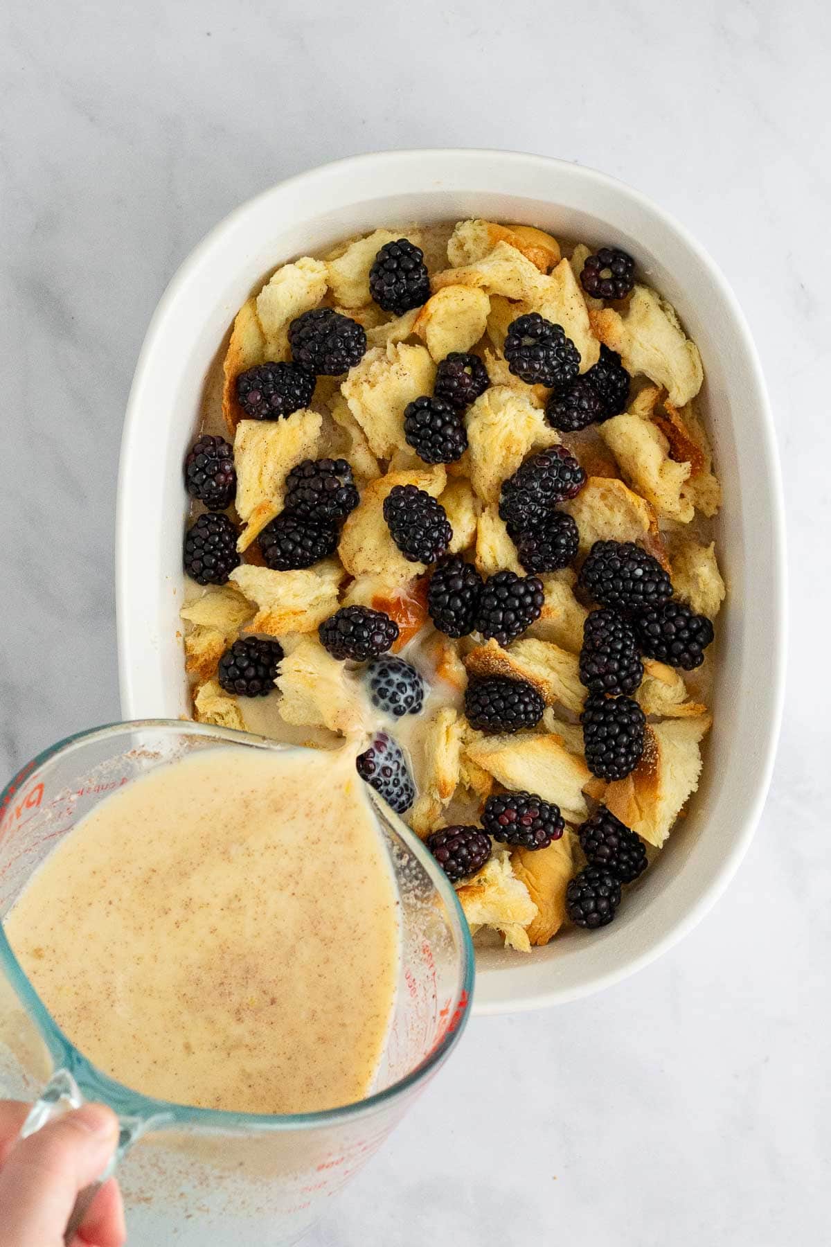 A hand pouring custard over ingredients in a baking dish for Blackberry Bread Pudding.