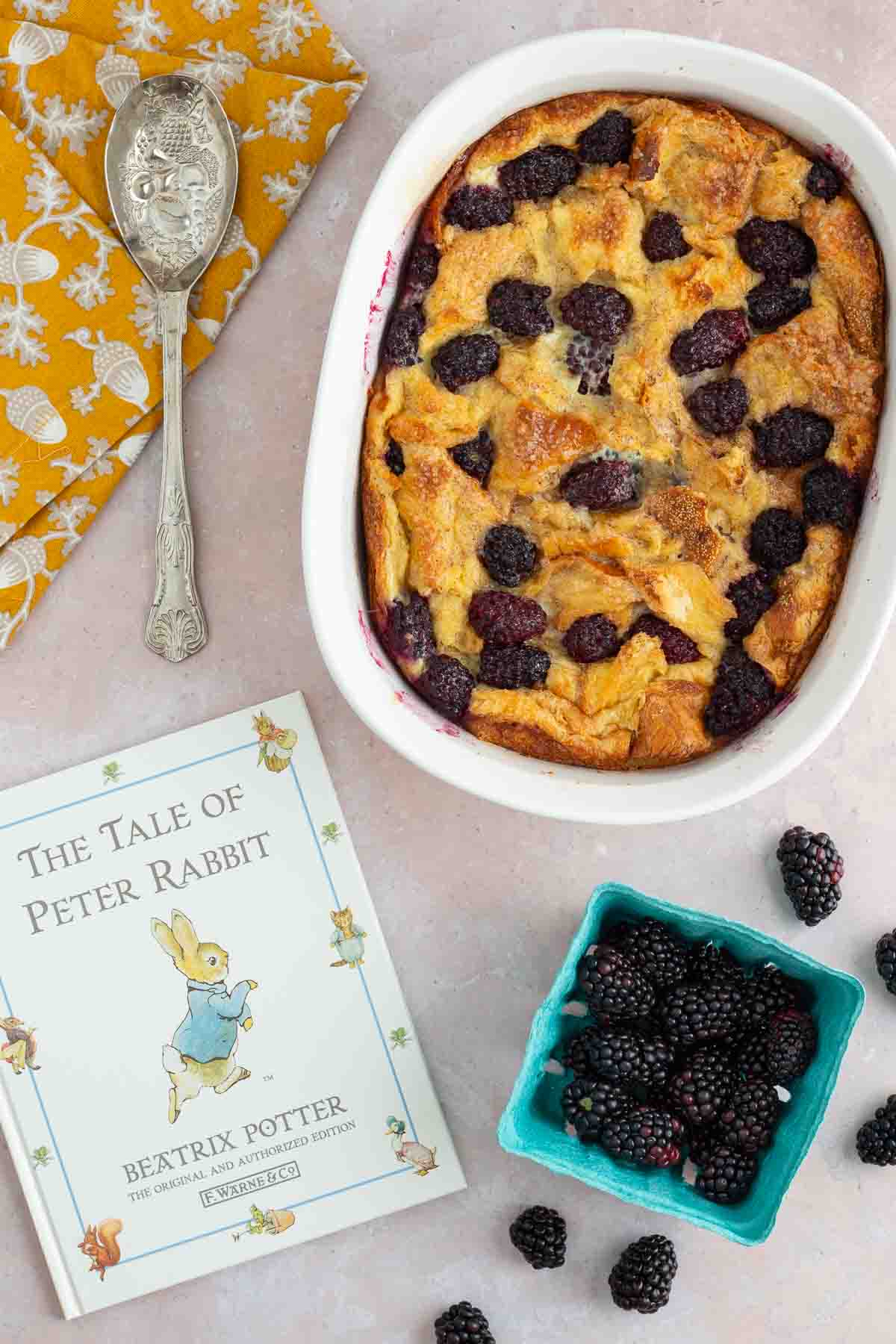 A baking dish of blackberry bread pudding with the book The Tale of Peter Rabbit next to it, along with fresh blackberries and a spoon.