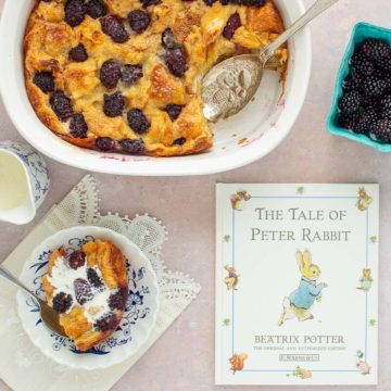 A baking dish of Blackberry Bread Pudding with the book The Tale of Peter Rabbit next to it, along with a serving in a bowl and a container of blackberries.