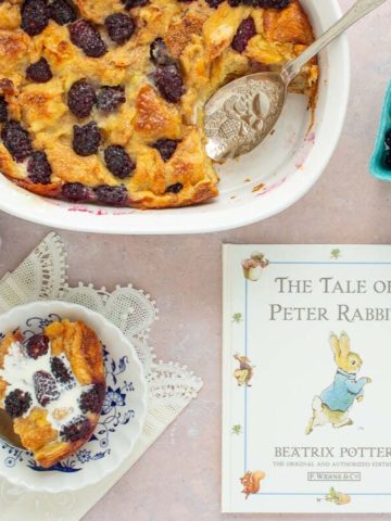 A baking dish of Blackberry Bread Pudding with the book The Tale of Peter Rabbit next to it, along with a serving in a bowl and a container of blackberries.