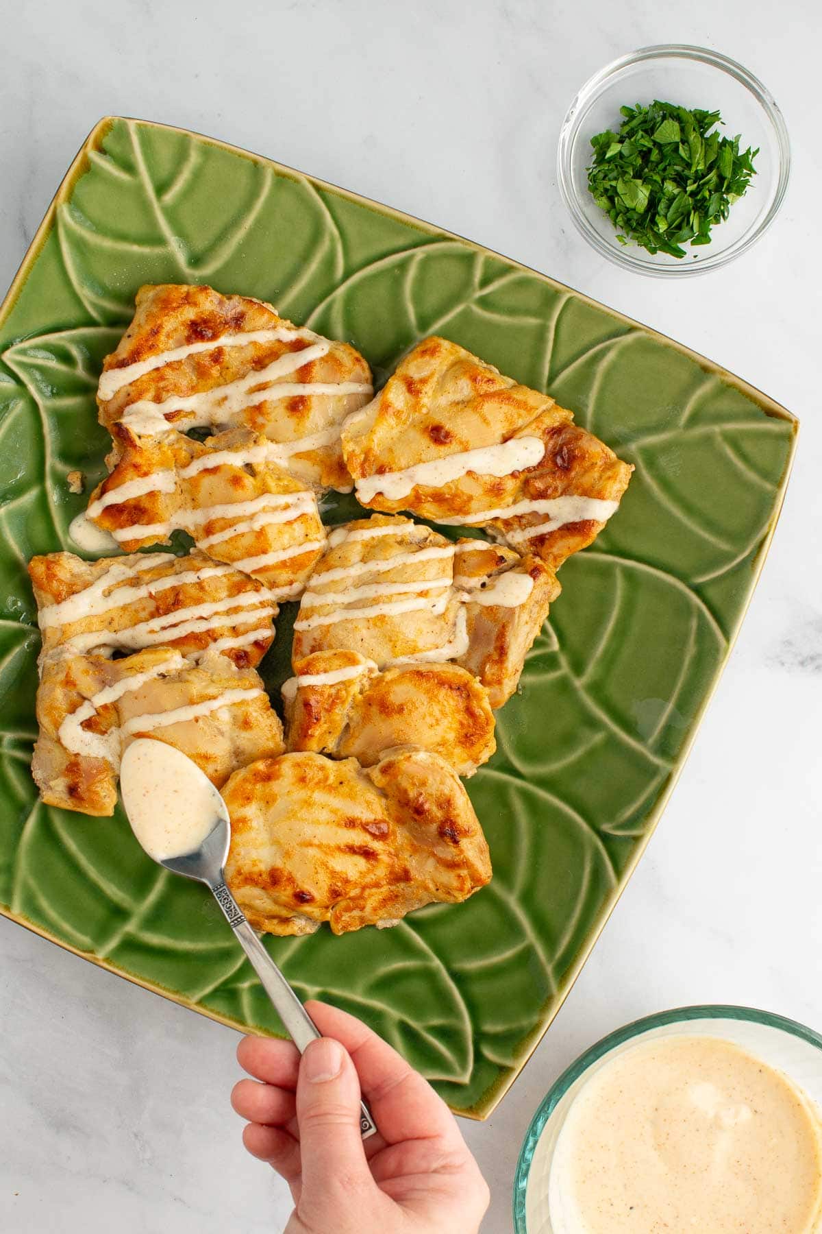 A hand drizzling yogurt sauce over baked chicken thighs on a platter.