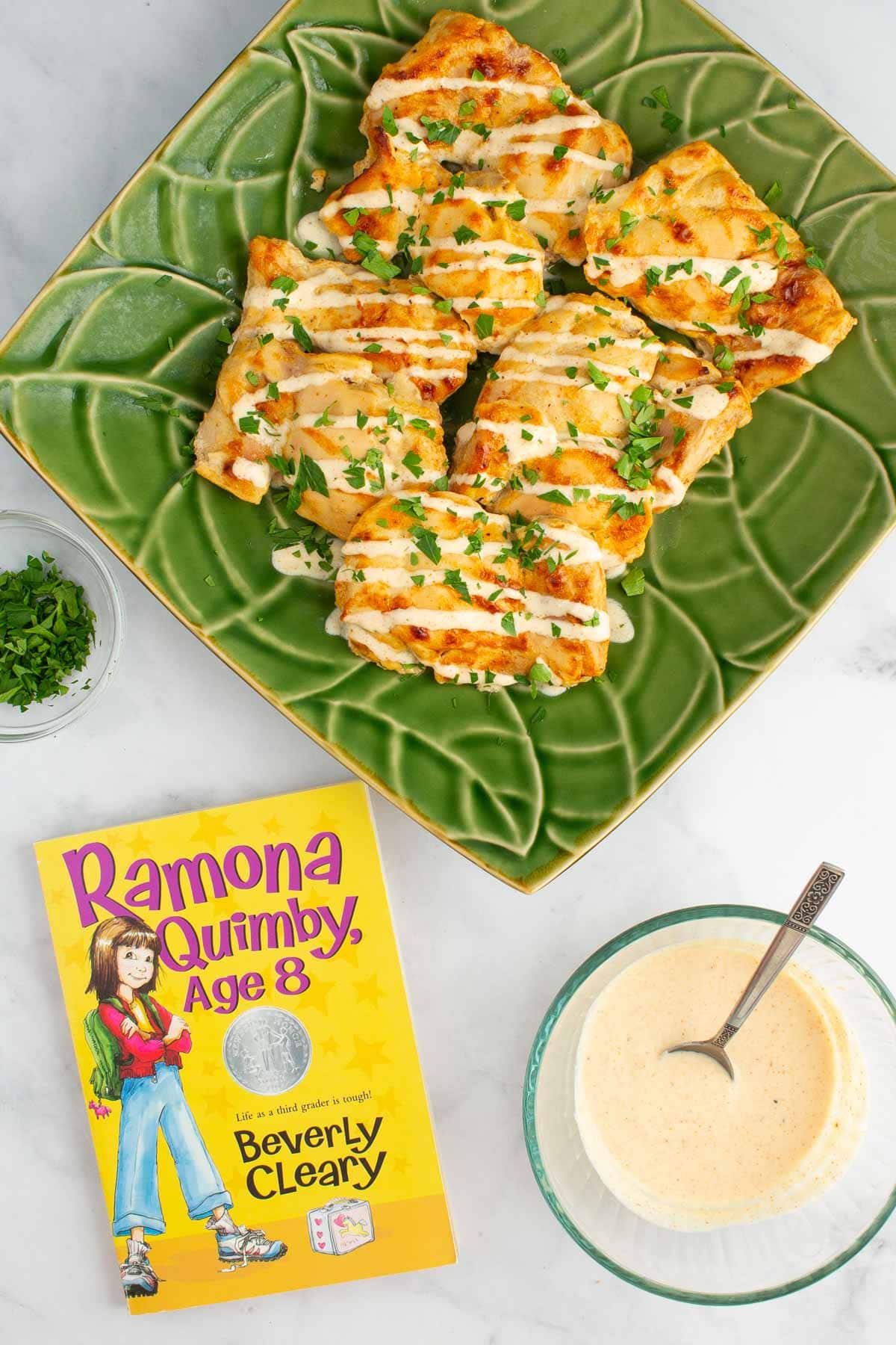 A platter of Juicy Yogurt-Baked Chicken with bowls of parsley and yogurt sauce nearby, and the cover of the book "Ramona Quimby, Age 8"