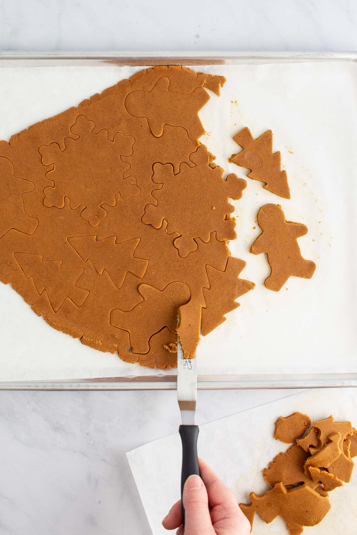 A hand using an offset spatula to remove dough scraps from cut-out easy gingerbread cookies.